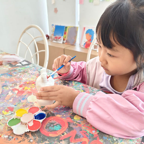 Child painting a ceramic object with a brush on a table with art supplies.